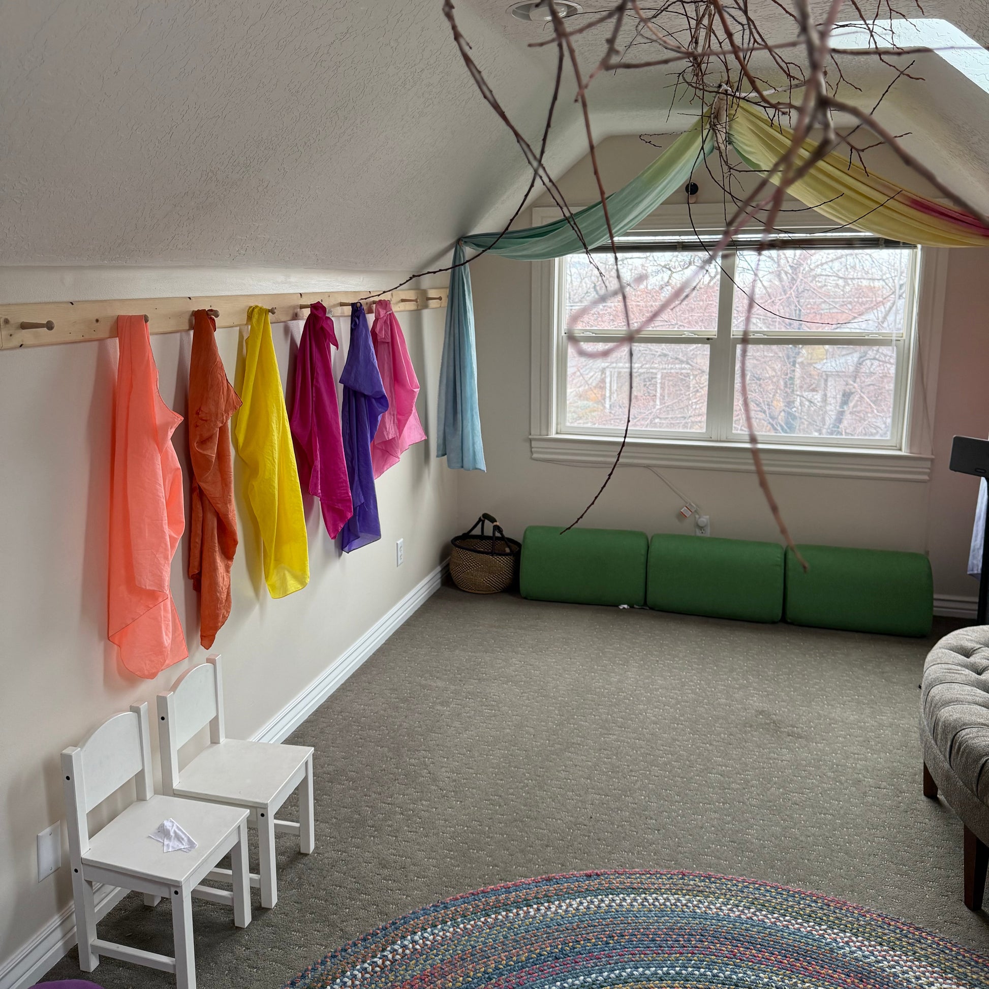 A child's playroom or attic space with a slanted ceiling, featuring colorful fabric scarves draped from a wooden beam, bright clothing hanging on a wall, green foam cushions by a window, and a round woven rug in the foreground.