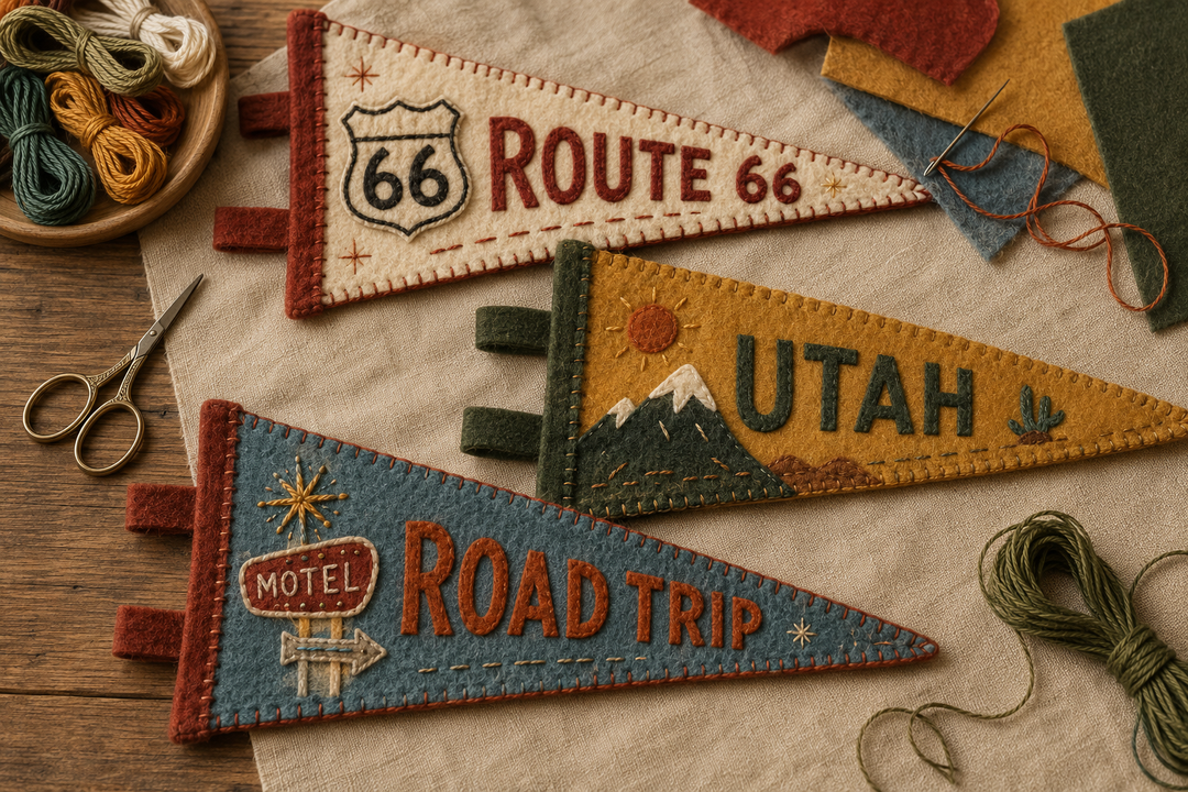 Three handcrafted felt pennant flags with vintage travel motifs are arranged on a fabric surface with sewing supplies nearby. The pennants read "Route 66," "Utah," and "Motel Road Trip," with embroidered mountains, stars, and decorative stitching.
