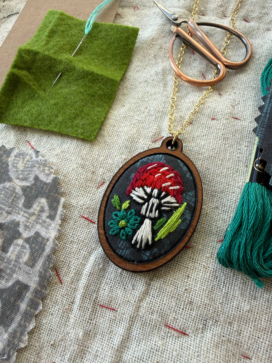 Embroidered pendant with floral design on a textured fabric background with embroidery tools.