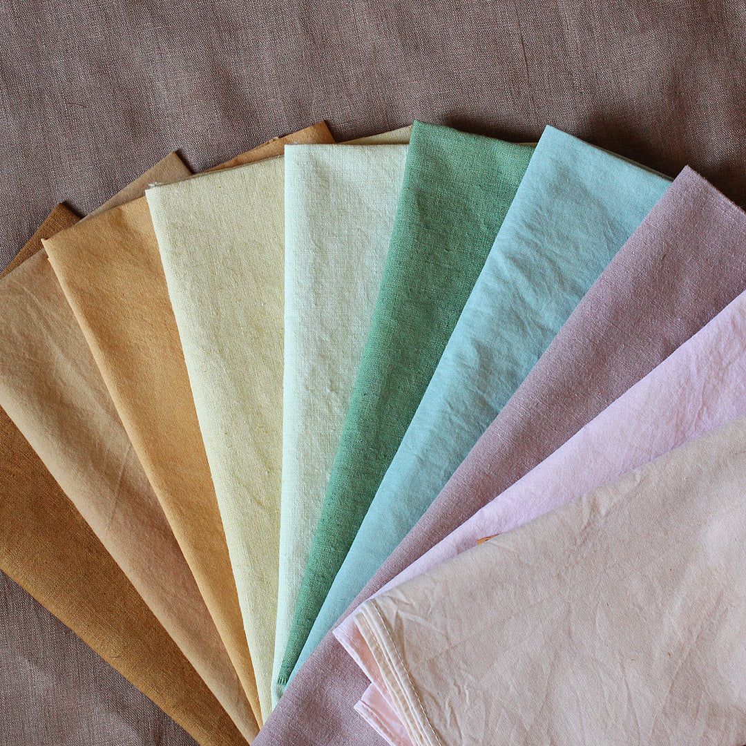 Assorted naturally dyed fabric swatches displayed in a fan arrangement, showcasing a range of colors including beige, green, blue, pink, and purple.