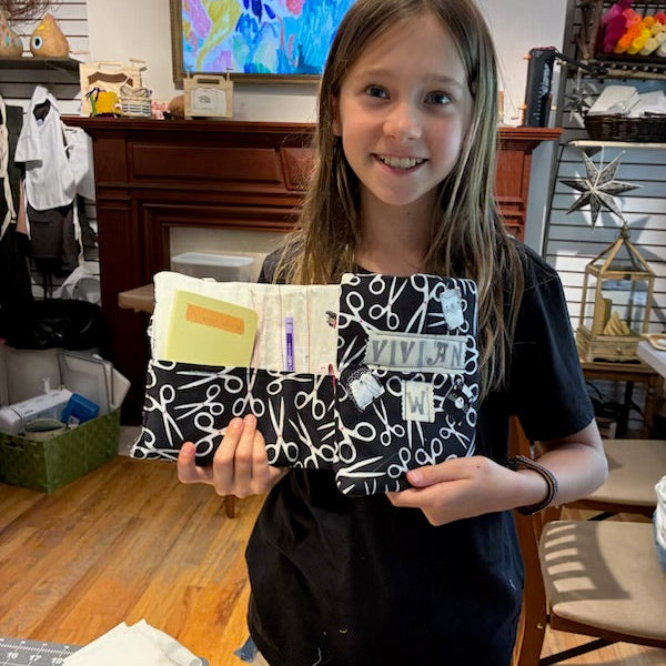 A young girl with long light brown hair smiles while holding a black and white patterned journal or notebook with several attached paper tabs in a home setting with wooden floors and furniture visible.