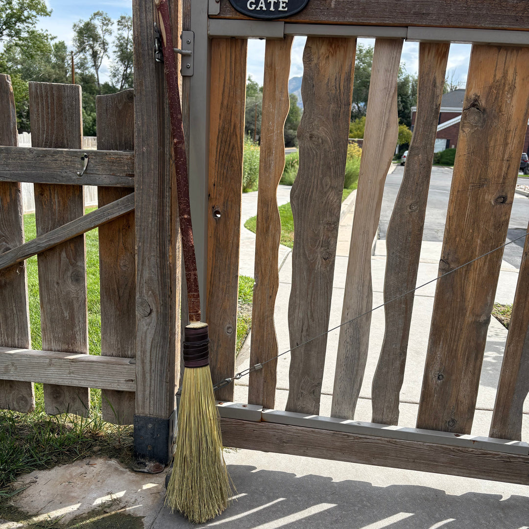 A rustic straw broom with a long brown wooden handle leans against a weathered wooden gate outdoors. A small black sign above the gate reads &