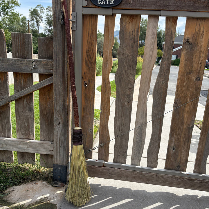 A rustic straw broom with a long brown wooden handle leans against a weathered wooden gate outdoors. A small black sign above the gate reads &