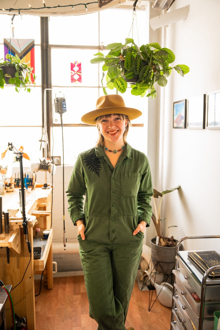 Person wearing a green jumpsuit and hat in a room with plants and furniture.