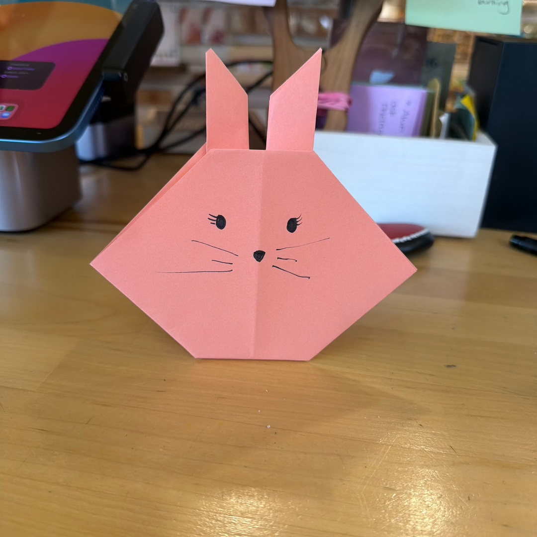 A pink origami paper bunny with simple black hand-drawn eyes, nose, and whiskers, standing on a wooden desk with office items in the blurred background.