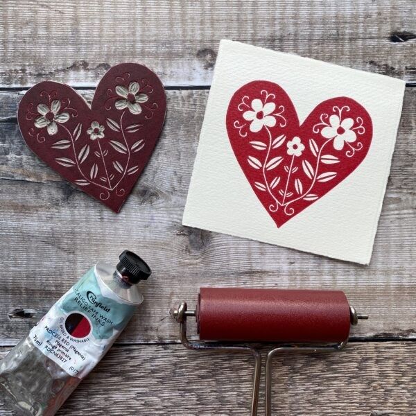 Heart-shaped stamp with floral design on a wooden surface, next to a printed card and printing tools.
