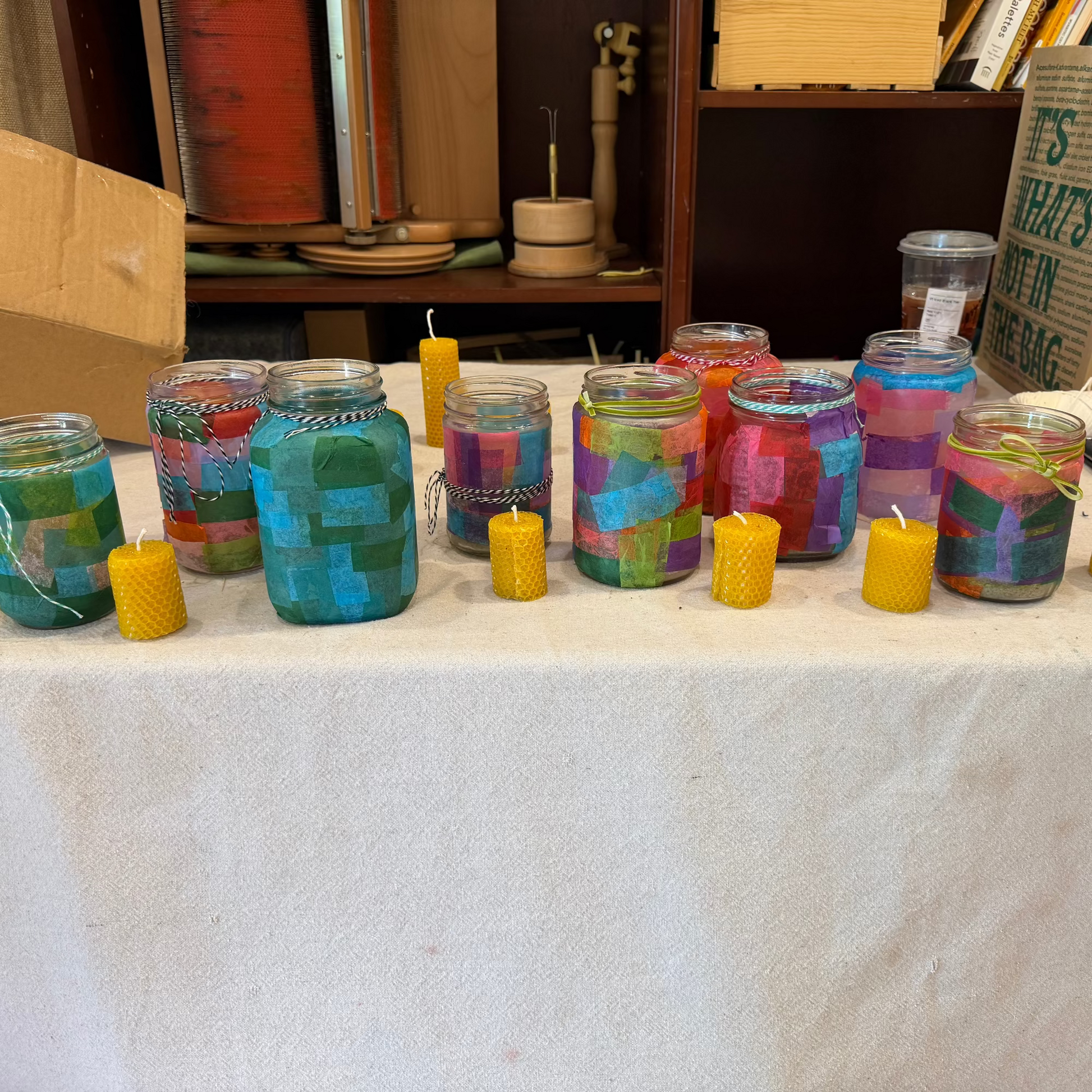 A display of handmade mason jar candles with colorful mosaic-patterned wax and small yellow pillar candles, set on a white tablecloth with a workshop background.