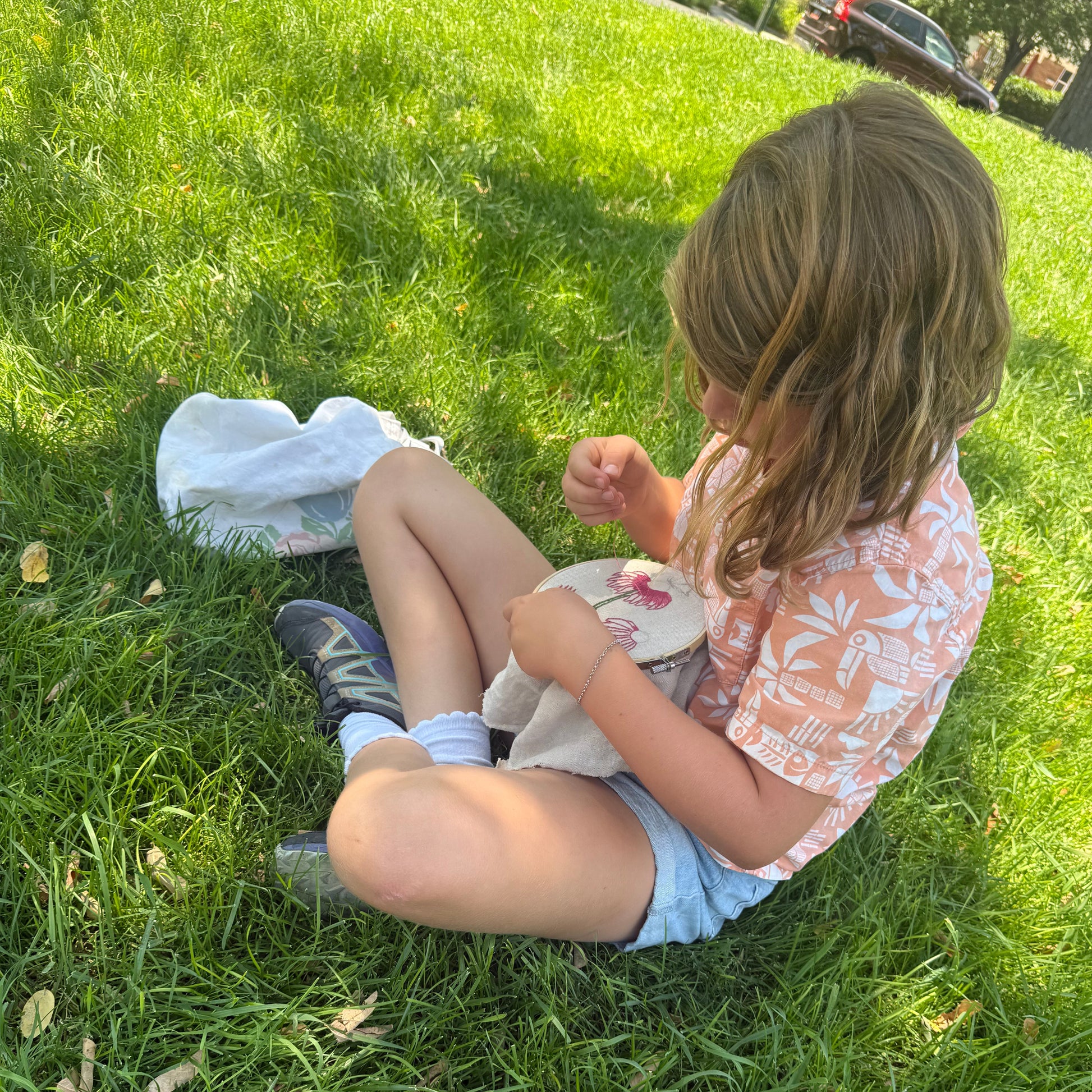 A young girl with blonde hair sits on a green grass lawn, wearing a peach and white patterned t-shirt, light blue denim shorts, and dark athletic shoes. She is holding a small white plush toy or bag.