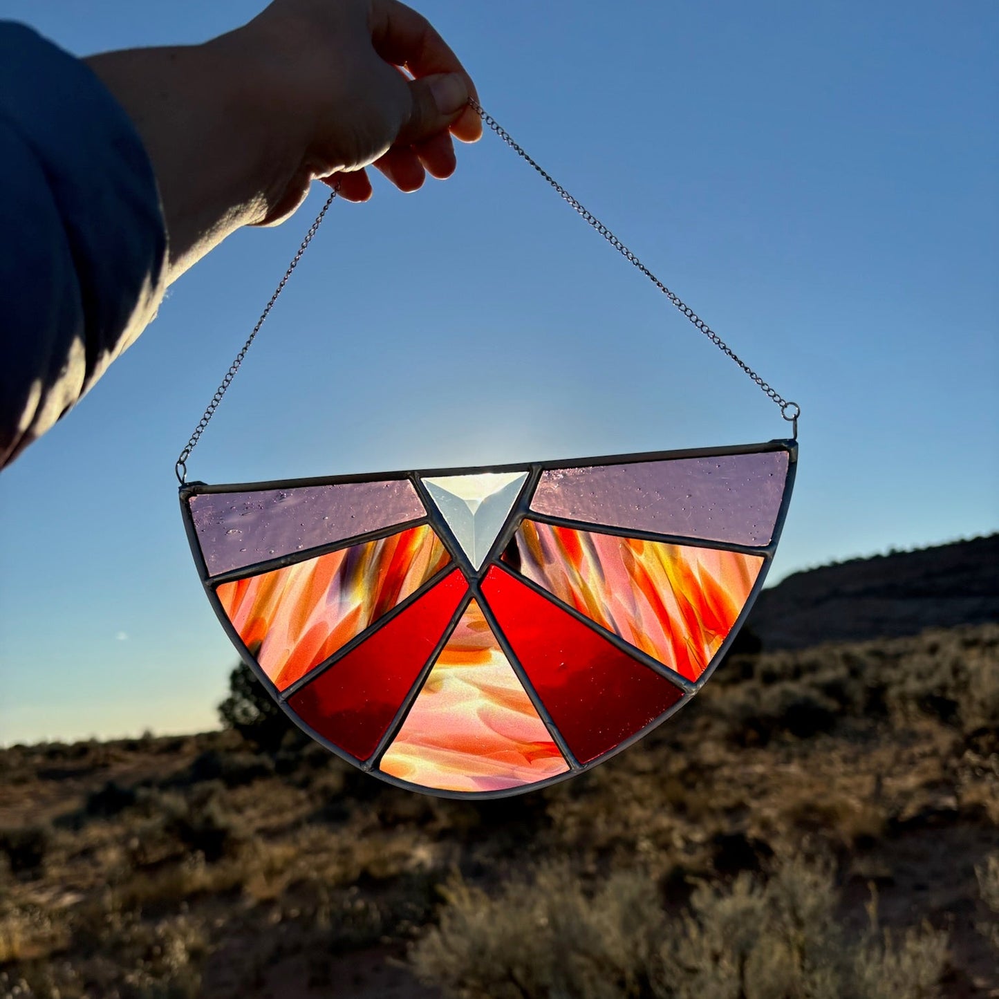 A hand holds a stained glass suncatcher in front of a desert landscape at sunset. The suncatcher features a semi-circular design with red, orange, yellow, and purple stained glass panels separated by dark metal lines, suspended by a thin metal chain.