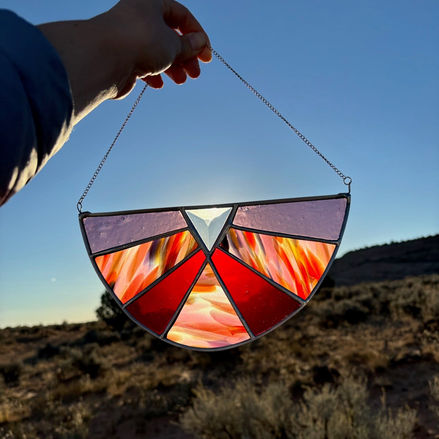 A hand holds a stained glass suncatcher in front of a desert landscape at sunset. The suncatcher features a semi-circular design with red, orange, yellow, and purple stained glass panels separated by dark metal lines, suspended by a thin metal chain.