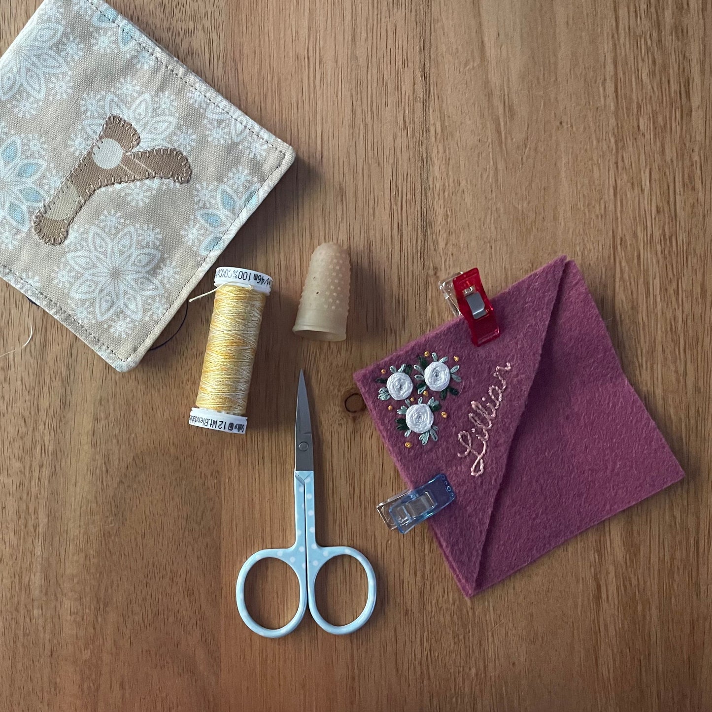 A styled flat-lay of sewing and crafting supplies on a wooden tabletop, including a beige fabric pouch with a blue floral pattern and brown bird motif, a spool of yellow thread, a beige thimble, a pair of silver scissors, and a purple felt pocket square with embroidered text and buttons.