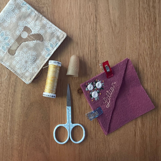 A styled flat-lay of sewing and crafting supplies on a wooden tabletop, including a beige fabric pouch with a blue floral pattern and brown bird motif, a spool of yellow thread, a beige thimble, a pair of silver scissors, and a purple felt pocket square with embroidered text and buttons.