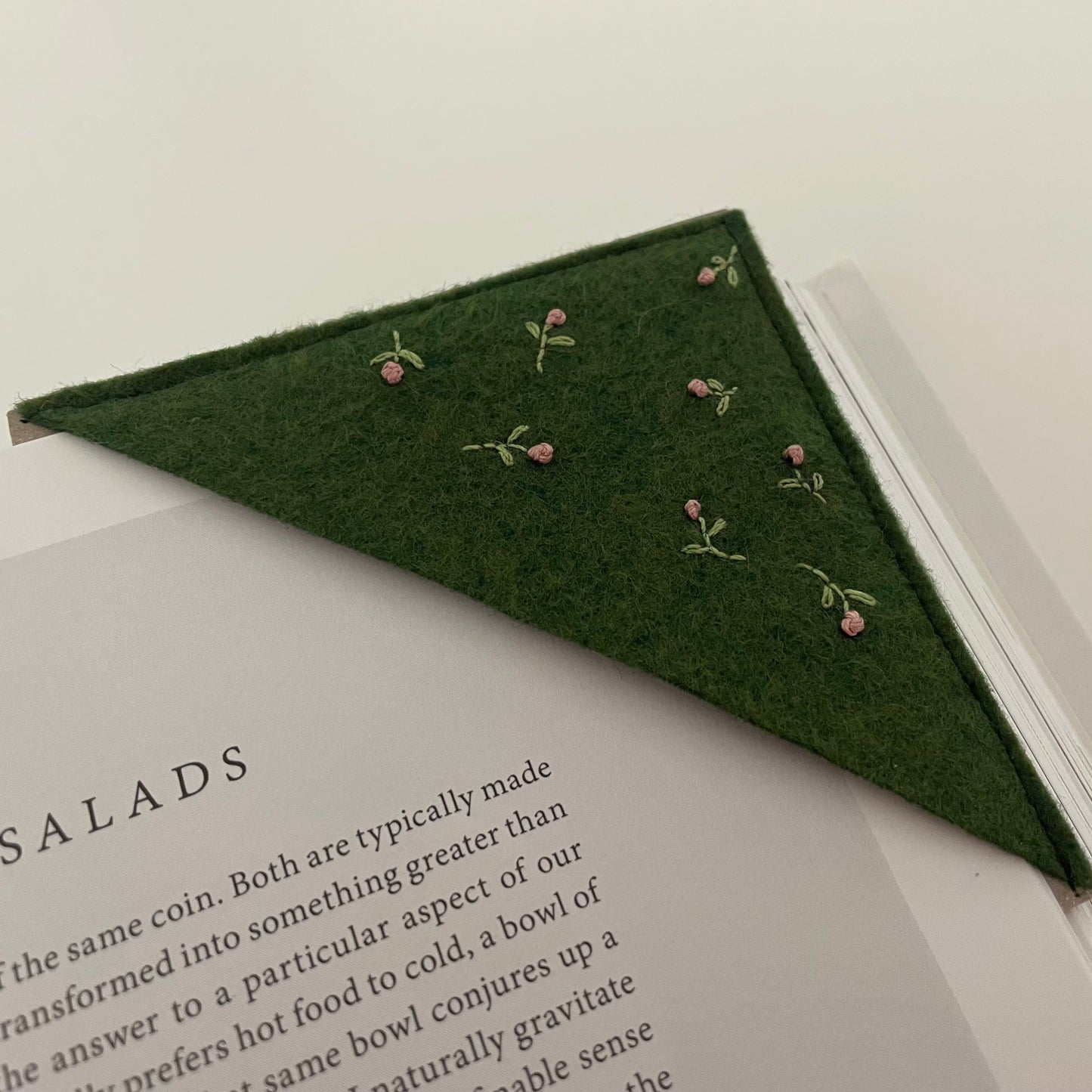 A triangular dark green felt bookmark with small embroidered pink flowers and green stems, placed on an open book.