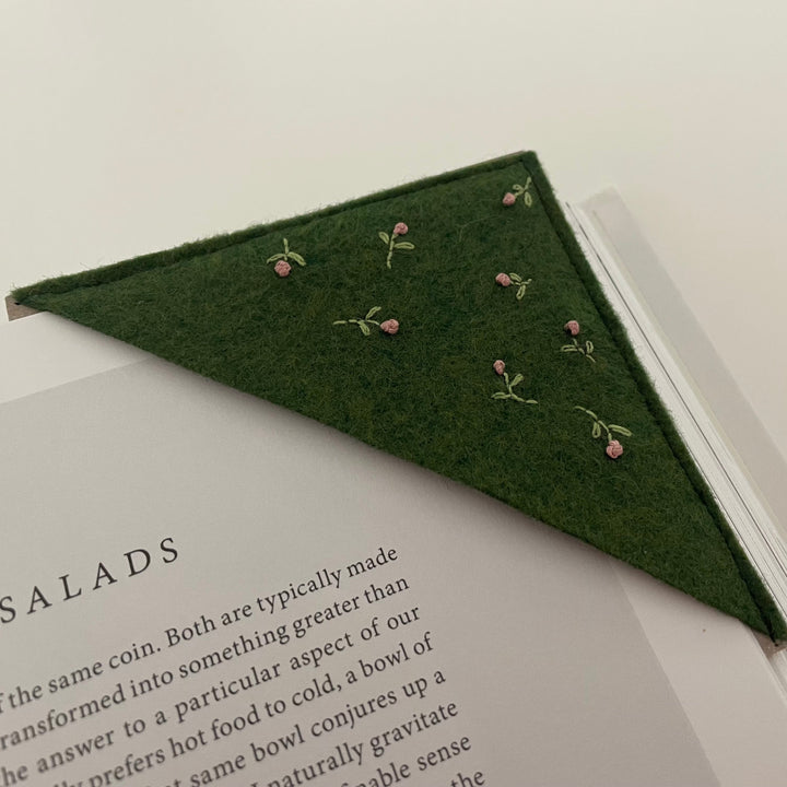 A triangular dark green felt bookmark with small embroidered pink flowers and green stems, placed on an open book.