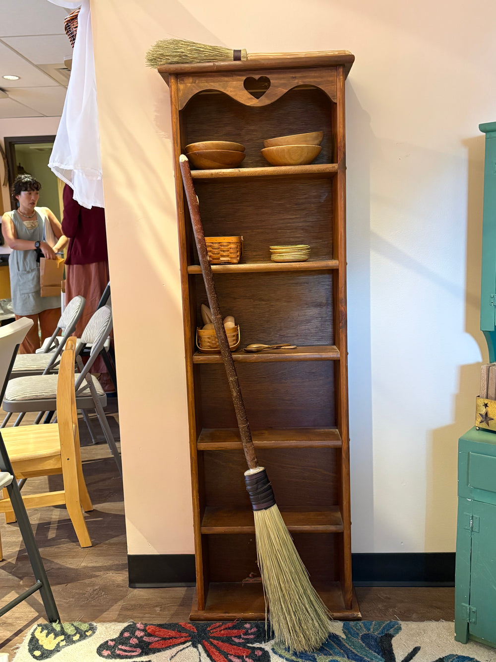 A tall rustic wooden shelving unit with five open shelves, styled with bowls, baskets, and a small heart-shaped ornament at the top. A long-handled straw broom leans against the unit on the floor in an indoor room with chairs and a wall visible.