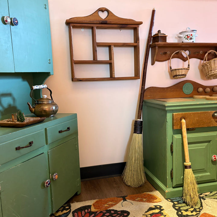 A styled vintage play kitchen corner featuring a sage green toy stove and matching toy cabinets, complete with a wooden wall shelf, hanging baskets, a metal kettle, and a butterfly-print rug on a light pink wall.