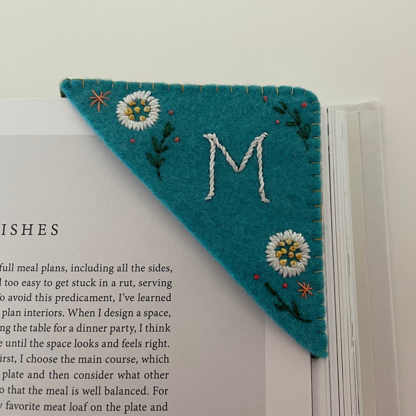 A teal felt book bookmark with a white embroidered letter 'M' and daisy flowers, shown bookmarking an open book.