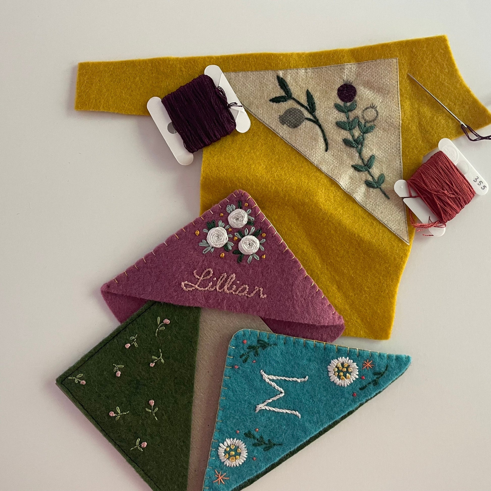 A collection of handmade felt accessories laid out on a white surface, including a mustard yellow belt with a triangular embroidered fabric panel, a purple felt tag with the name 'Lilian' in gold script, a green felt tag with small floral embroidery, and a teal felt tag with a white monogram 'M' and daisy embroidery. Two spools of thread and a needle are also visible.